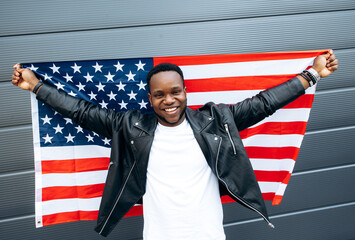Happy young handsome smiling black guy stands with hands up and holds US flag behind him, outdoors