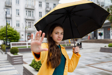 A young pretty teenage girl walks around the city in rainy weather. The girl dressed in colored clothes and rubber boots forbids the photographer to take photos