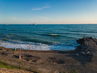 View of the Black Sea coast with people resting, lit by sunlight at sunset, with a breakwater and ships on the horizon against the blue sea on a sunny evening.