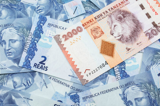A close up image of a two thousand Tanzanian shilling bank note on a background of Brazilian two reais bank notes