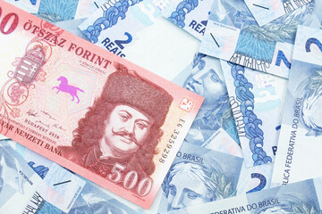 A close up image of a red Hungarian five hundred forint bank note with Brazilian two reais bank notes in the background