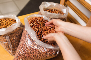 Woman with hands full with raw peanuts, taking it out of big transparent bag on wooden table. Nuts in bulk.