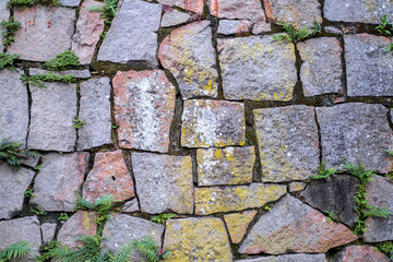 Wall of large gray stones partially covered with yellow and red moss and green plants between them. Paving stone road.