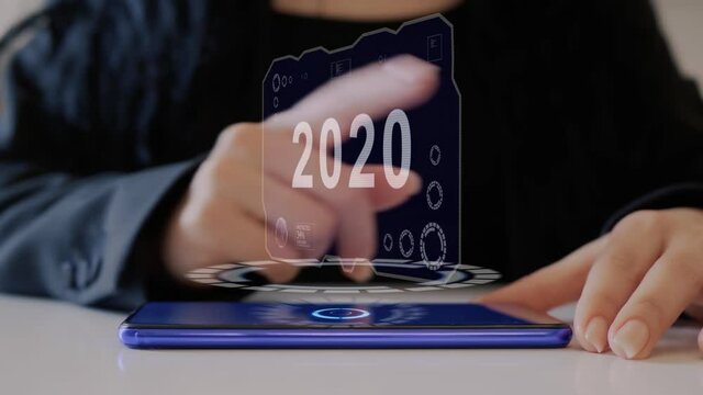 Female hand interacts HUD hologram with text 2020. Woman in black uses conceptual holographic technology of the future on the screen of a smartphone lying on a table
