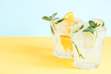 Two glass with lemonade or mojito cocktail with orange, mint and ice over yellow background with copy space.