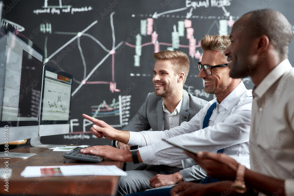 Wall mural Dreams Come True. Side view of three colleagues traders sitting by desks in front of computer monitors and discussing strategies. Blackboard full of charts and data analyses in background