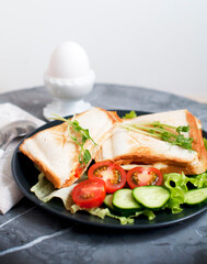 Hot sandwiches with cream cheese, cucumber, ham, tomatoes and peas sprouts on black plate. Breakfast with egg mist on white stand. Top view