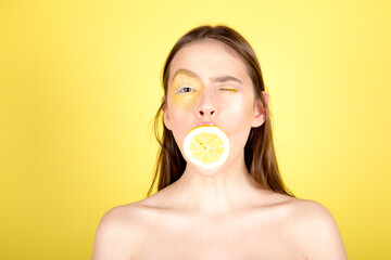 Beautiful young woman with wet make up holding juicy lemon slice in front of eyes on yellow background. Perfect Fresh Skin. Healthy Lifestyle.
