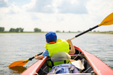 Fototapeta premium A little boy kayaks on the river in the summer and rowing an oar. The boy is floating in a boat on the river.