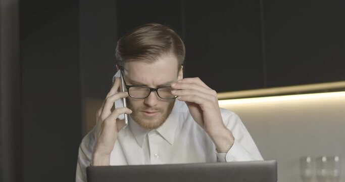 Slowmo close-up of confident man talking on the phone and looking at laptop screen. Portrait of serious concentrated Caucasian businessman discussing project on smartphone. Cinema 4k ProRes HQ.