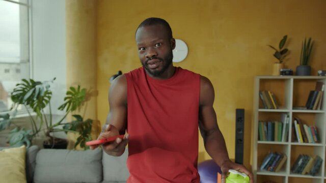 Portrait of funny afro-american athletic man with smartphone looking baffled into camera while reading social media messages. Communication. Home indoors.