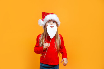 Fototapeta na wymiar Funny child girl in Santa red hat holding paper beard on blue background.