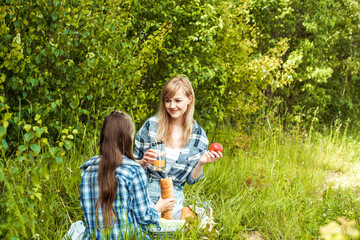 young mother and little daughters in bright sweaters on a picnic in nature indulge in apples.Picnic in the forest, green park, wicker basket, fruit, juice and white bread. Outdoors.