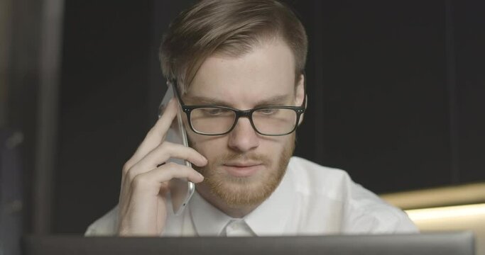 Worried confident man in eyeglasses talking on the phone and looking at laptop screen. Close-up portrait of concentrated Caucasian businessman having business problems. Cinema 4k ProRes HQ.