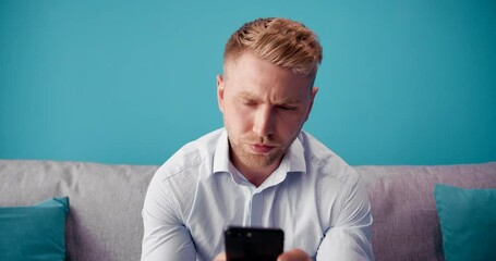 Young bearded guy with stylish haircut in formal clothing sitting on grey couch and using smartphone. Concept of modern technology and leisure time.