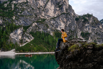 Obraz premium Brunet man in yellow raincoat sitting on the cliff and watching the boat floats on the lake Braies (Lago di Braies, Wildsee) on background of high mountain wall and cloudy sky in Dolomites Alps. Italy