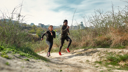 Preparing for a marathon. Young sporty african couple or friends in fitness clothing running uphill outdoors in nature or city park