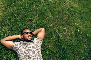 Cheerful attractive afro american guy student resting while lying on a grass in the park at the university. Space for text - Powered by Adobe