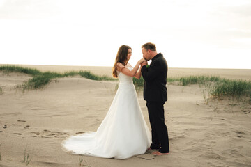 Fototapeta na wymiar Wedding couple standing and posing on sandy beach. Groom kissing bride's hands. Barefoot husband. Wedding day concept.