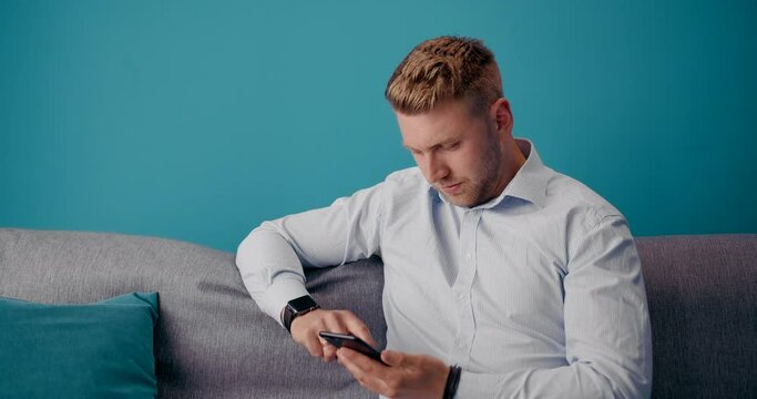 Smiling young man in blue shirt relaxing on grey couch and using personal smartphone. Bearded guy enjoying free time at home with digital gadget.