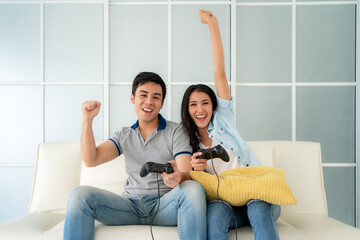 Asian couple man ans woman to playing video games with joysticks while sitting in sofa in living...