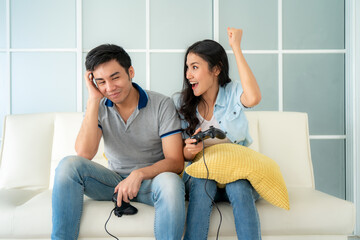 Obraz premium Asian couple man ans woman to playing video games with joysticks while sitting in sofa in living room at home and woman winning in game looking man lose.