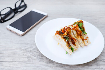 Grilled chicken sandwiches in a plate, smartphone, and eyeglasses on wooden table