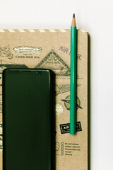 The notebook is on a wooden table, with a cell phone and a green pencil on it.