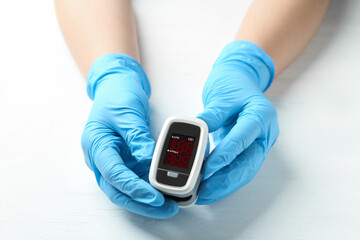 Doctor in gloves holding fingertip pulse oximeter at white wooden table, closeup
