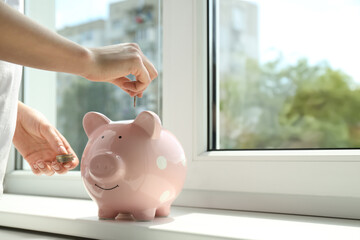 Woman putting money into piggy bank at window sill indoors, closeup. Space for text