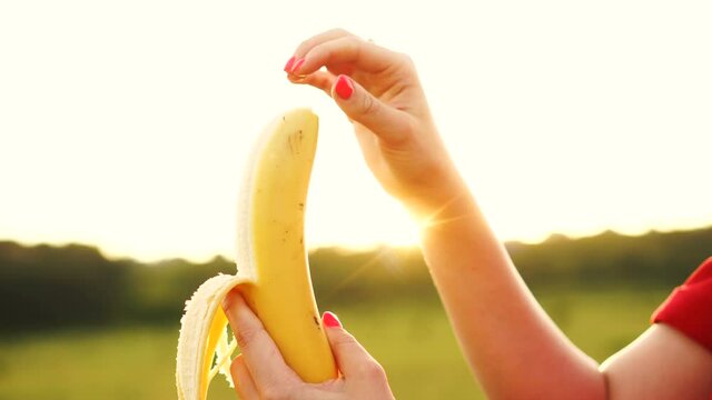 Woman peeling and biting the banana. Ripe yammy banana in female hands on sunset background