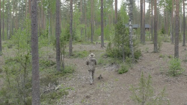 Man in camouflage walking in the woods near a forest house.