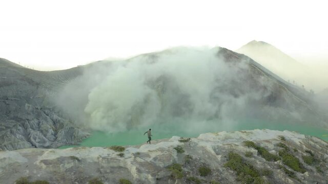Aerial panning shot of man running on rocky mountain peak near lake, drone flying over natural landscape with smoke - East Java, Indonesia