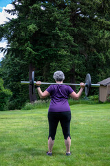 Middle aged caucasian woman with gray hair lifting a barbell with black plates, fitness outside on the lawn
