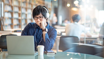 Smart Asian business man with glasses and headphones listening to music while using laptop computer in public library. Male freelance working from coworking space. Internet of things concept.
