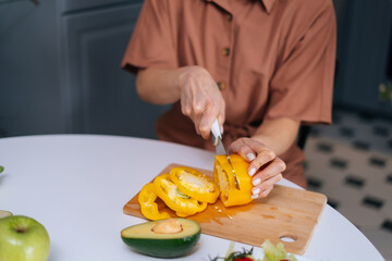 Close-up of woman hands cutting fresh bell pepper using knife on wooden cutting board. Young woman cutting yellow bell pepper with a knife for vegetable salad. Concept of healthy eating.