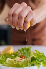 Close-up of women's hands squeezing juice from fresh yellow lemon into salad bowl with sliced vegetables. Red bell pepper, avocado, lemon on the table. Concept of healthy food lifestyle.
