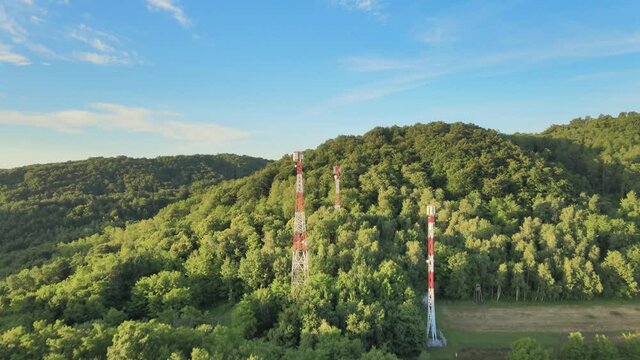 Backward moving aerial over dense green mountains with Five G telecommunication towers standing tall on bright sunny day.