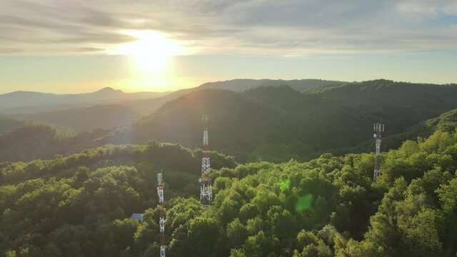 Rotating aerial view of 5G telecommunication towers standing tall on dense green mountains against beautiful sunlight