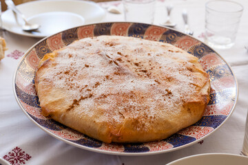 Food shot of a family size crispy delicious gourmet moroccan pastilla on an ornaments plate covered with icing sugar and cinnamon on a linens tablecloth with glasses, knife, fork and napkins.  