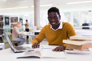 Fototapeta na wymiar Portrait of smiling african-american male student with laptop and book in public library