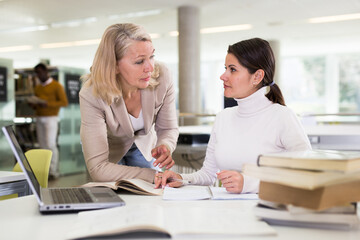 Friendly female tutor helping to diligent positive girl preparing for exam in library
