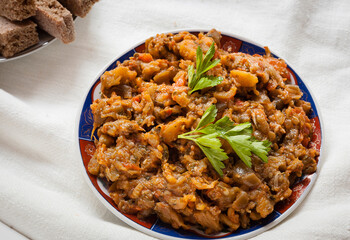 Top view food shot of traditional Moroccan mashed aubergine and tomato with spices salad Zaalouk plate with parsley leaves and homemade rye bread plated on a white tablecloth linens.