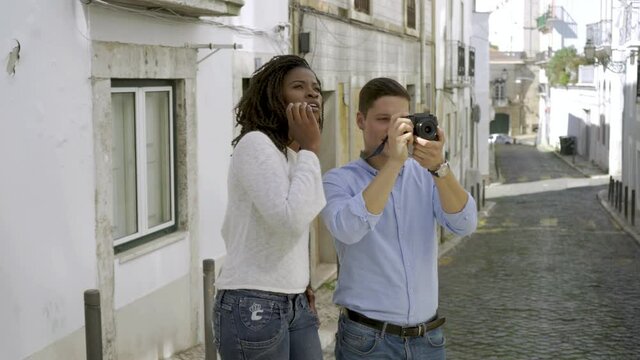 Multiethnic couple of tourists standing in old city street, holding camera, taking pictures of landmarks, laughing. Medium shot. Travel and photo concept