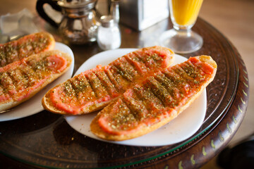 
Plate of toasted baguette bread with tomato sauce, Spanish olive oil, spices and sesame seeds on a white plate with orange juice, salt shaker and moroccan teapot. Mediterranean breakfast concept.
D