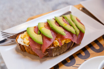 Complete healthy breakfast toasted bread with scrambled eggs, cheese, pastramy ham and avocado slices on a white plate with orange juices and cutlery on the woden background table. 