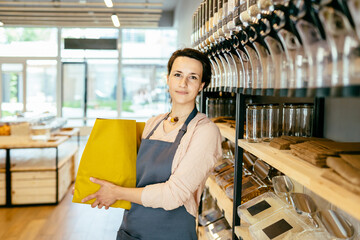 Portrait of a happy young positive brunette female assistant merchant in grey apron standing with paper bag with healthy food in store over rack counter service, consulting, helping, smiling.