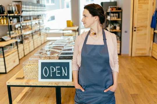 Mixed Race Woman Worker Or Store Owner With Standing In The Doorway Of Her Grocery Shop Looking Aside Smiling. Portrait Of Girl Assistant Wearing Apron Welcoming Inviting Open Door Of Store.