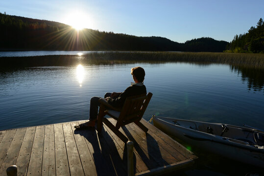 Man sitting alone on a wooden chair on a wooden dock/pier/jetty on a lake with kayaks around and the sun shining in the sky and reflected in the water