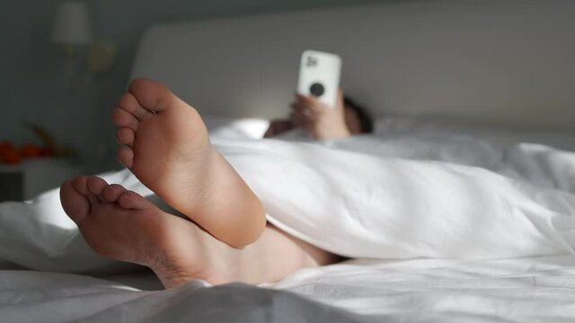 Female bare feet under white blanket, woman holding mobile phone lying in bed.